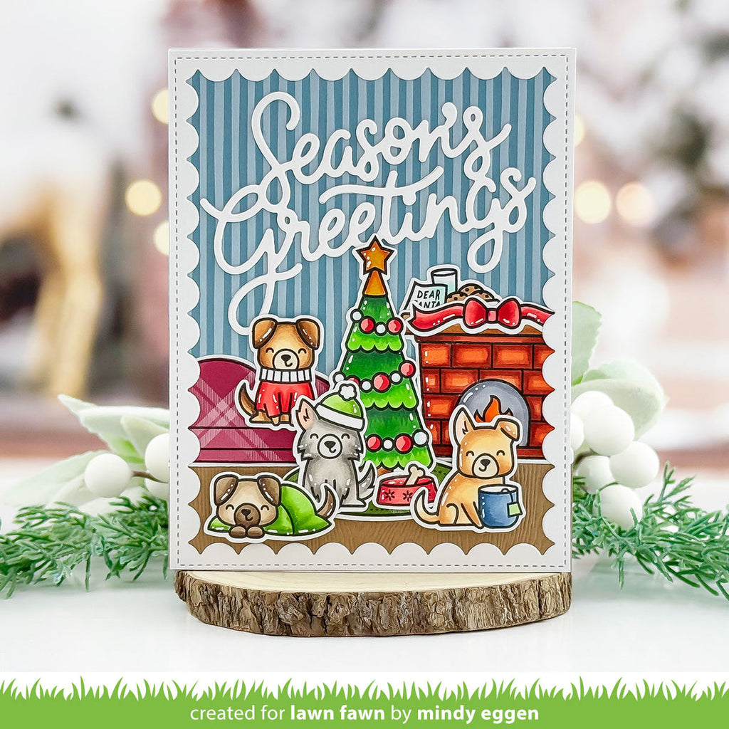 A handmade holiday greeting card featuring cartoon dogs around a Christmas tree and fireplace, displayed on a wood slice stand with blurred festive greenery in the background.