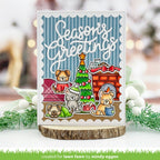 A handmade holiday greeting card featuring cartoon dogs around a Christmas tree and fireplace, displayed on a wood slice stand with blurred festive greenery in the background.