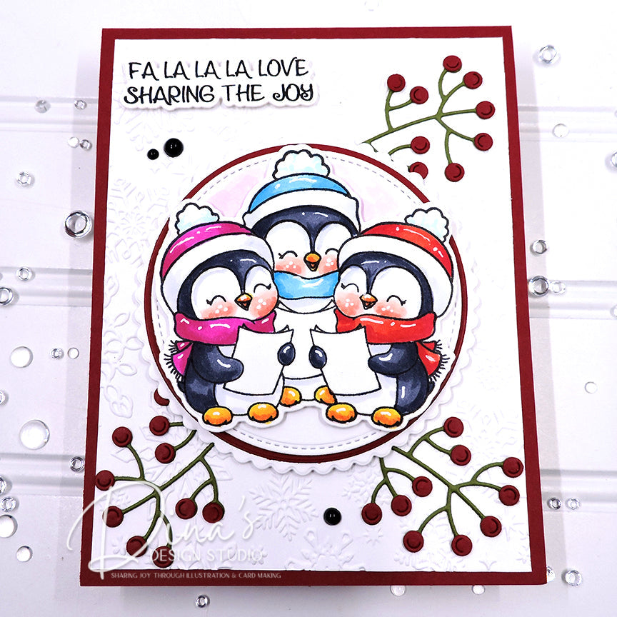 A handmade holiday greeting card featuring three penguins in winter hats and scarves singing under a banner that reads 'FA LA LA LA LOVE SHARING THE JOY'. The card is decorated with red berries and green branches, set against a white background with scattered clear rhinestones.