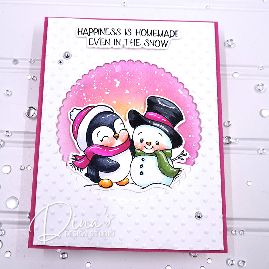 A handmade holiday greeting card featuring two cartoon penguins and a snowman under a pink scalloped circle, with the text 'HAPPINESS IS HOMEMADE EVEN IN THE SNOW' at the top. The card is photographed on a white textured surface with scattered rhinestones.