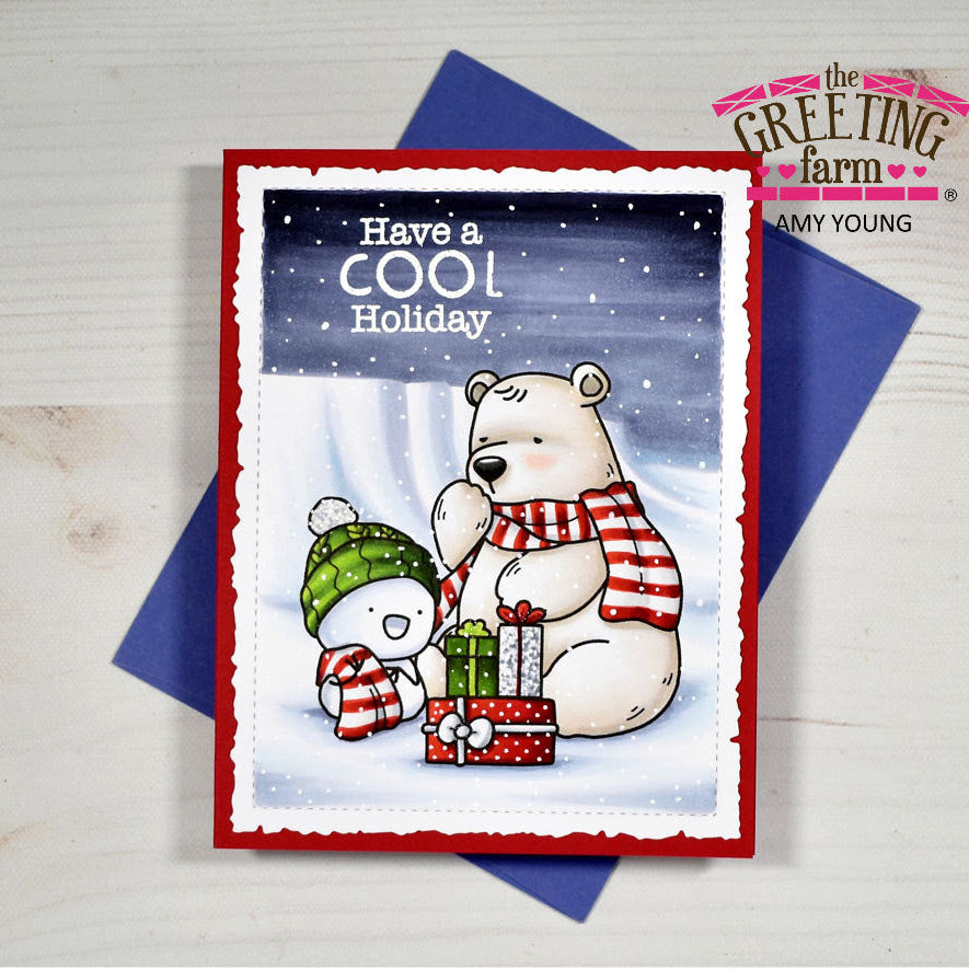 A handmade holiday greeting card from The Greeting Farm by Amy Young, featuring a cute cartoon polar bear and snowman with presents under a snowy night sky. The card is displayed on top of a blue envelope on a light wood background.
