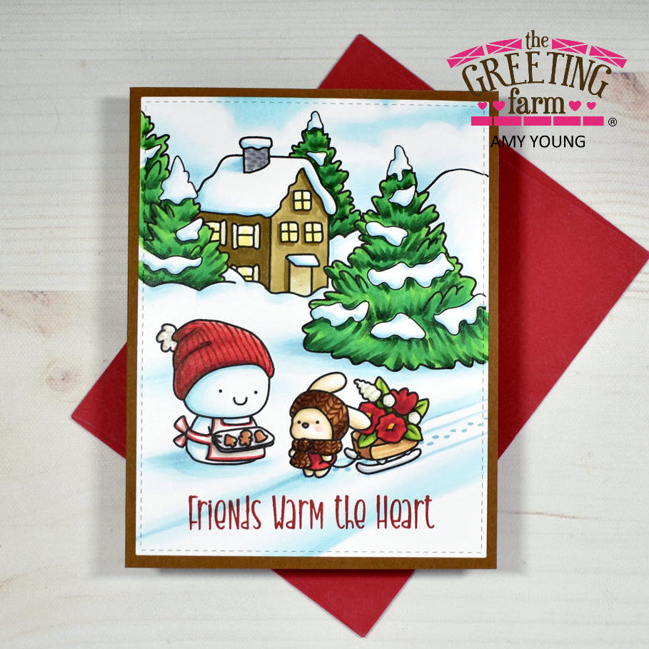 A handmade holiday greeting card from The Greeting Farm by Amy Young, featuring a winter scene with a snowman and a child on ice skates in front of a cozy house and evergreen trees. The card is displayed on top of a red envelope against a light wood background.