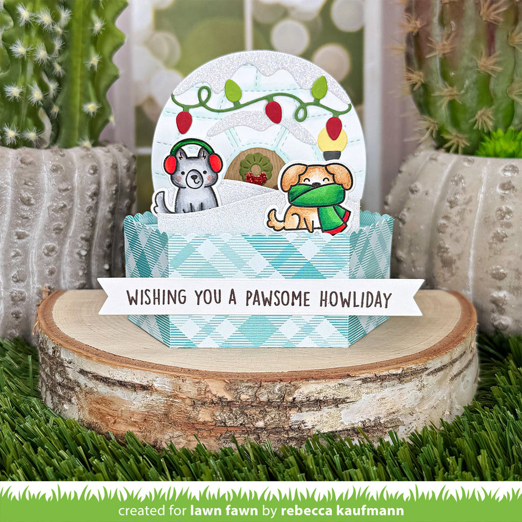 A handmade holiday greeting card or pop-up gift box display featuring two dogs in winter clothing under a snowy globe design, placed on a birch log with cacti in the background. A banner reads 'WISHING YOU A Pawsome Howliday' and a green credit line at the bottom says 'created for lawn fawn by rebecca kaufmann'.