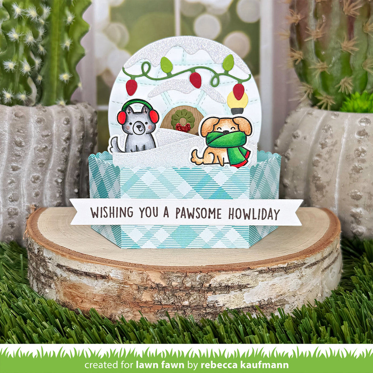 A handmade holiday greeting card or pop-up gift box display featuring two dogs in winter clothing under a snowy globe design, placed on a birch log with cacti in the background. A banner reads 'WISHING YOU A Pawsome Howliday' and a green credit line at the bottom says 'created for lawn fawn by rebecca kaufmann'.