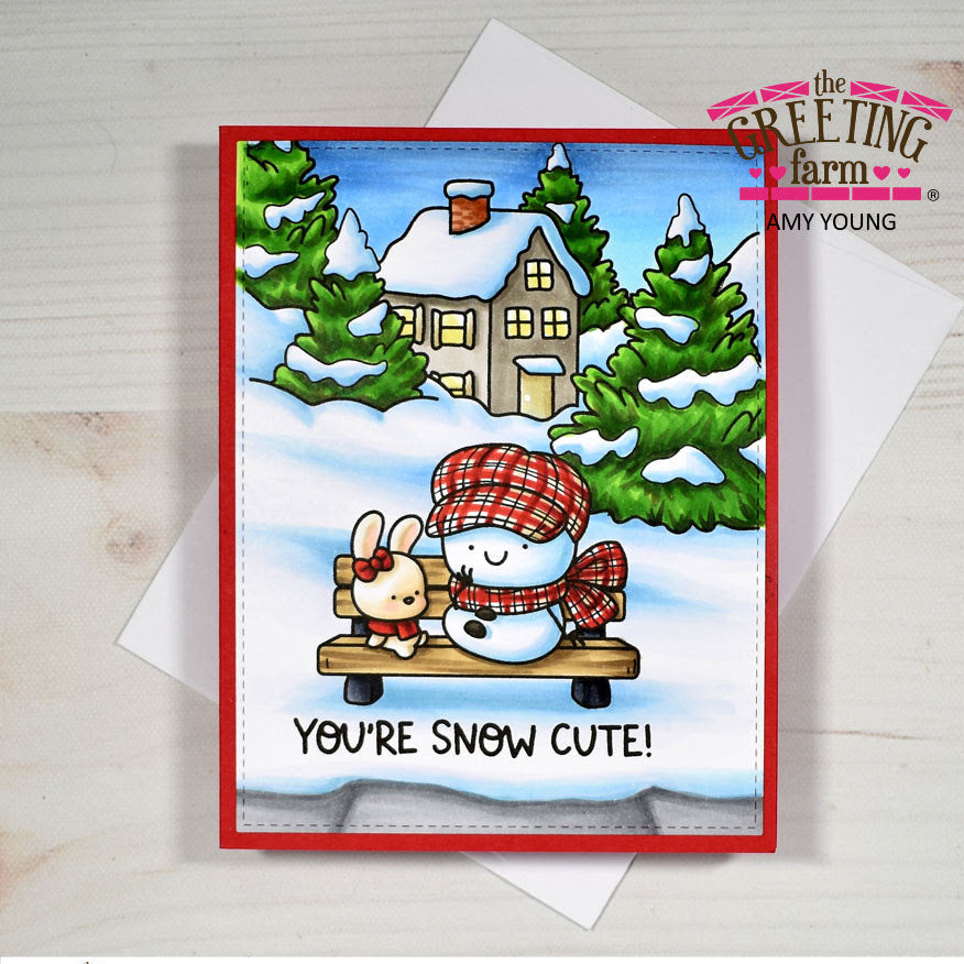 A handmade holiday greeting card with a red border, featuring a winter scene with a snowman and bunny on a bench in front of a snowy house and trees. The card is displayed on top of white envelopes on a light wood background, with 'The Greeting Farm' branding visible.