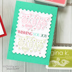 A handmade layered Christmas greeting card featuring a white scalloped stamp panel with hanging ornament and tree illustrations, the phrase “WISHING YOU JOY” in red and green, and a bright aqua green card base. The card is photographed on a light wood surface with partial ink pads visible around it.