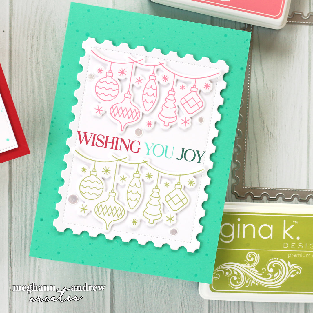 A handmade layered Christmas greeting card featuring a white scalloped stamp panel with hanging ornament and tree illustrations, the phrase “WISHING YOU JOY” in red and green, and a bright aqua green card base. The card is photographed on a light wood surface with partial ink pads visible around it.