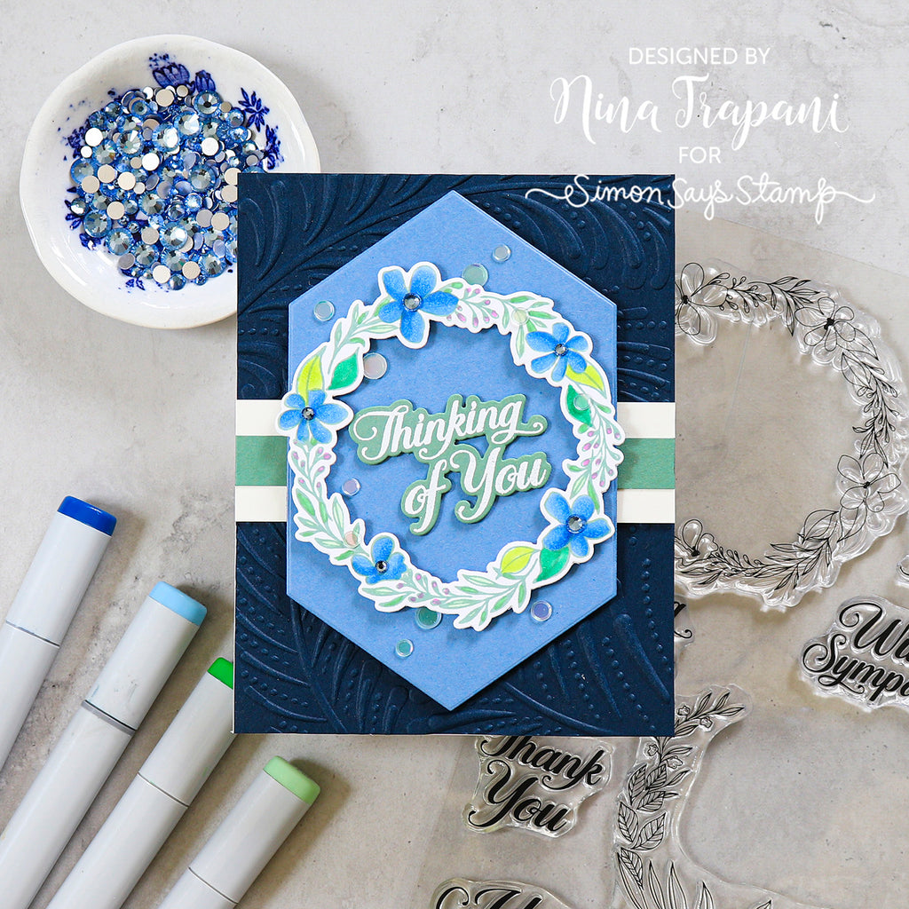 A handmade layered card featuring a blue hexagonal center with a floral wreath and the words 'Thinking of You,' styled on a light stone surface with markers, rhinestones, and clear stamps around it. Text in the upper right reads 'Designed by Nina Trapani for Simon Says Stamp.