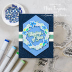 A handmade layered card featuring a blue hexagonal center with a floral wreath and the words 'Thinking of You,' styled on a light stone surface with markers, rhinestones, and clear stamps around it. Text in the upper right reads 'Designed by Nina Trapani for Simon Says Stamp.