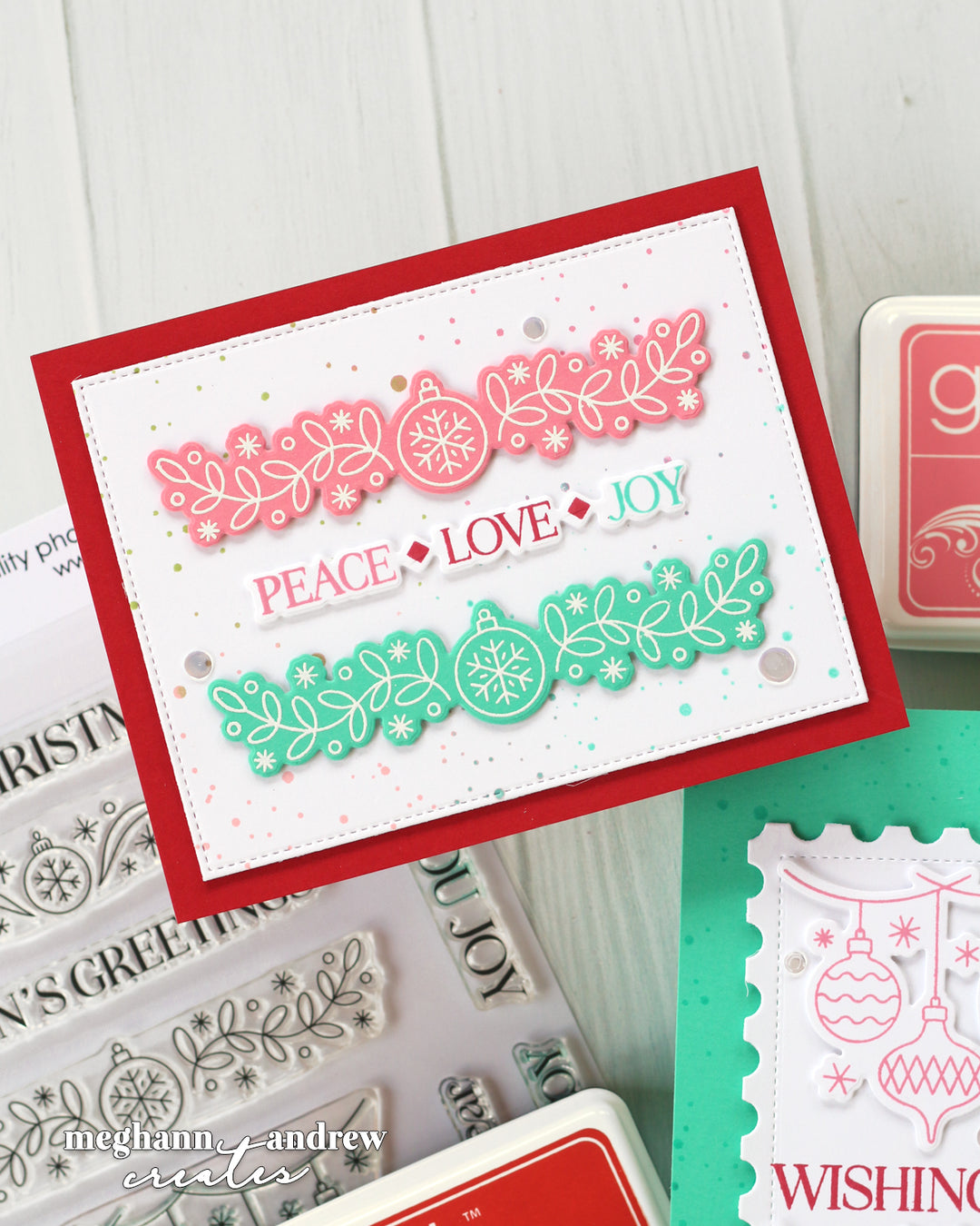 A handmade layered card featuring a red border, a pink holiday garland with snowflakes and ornaments, and a green garland with ornaments and snowflakes. The card has the words 'PEACE', 'LOVE', and 'JOY' layered in the center.