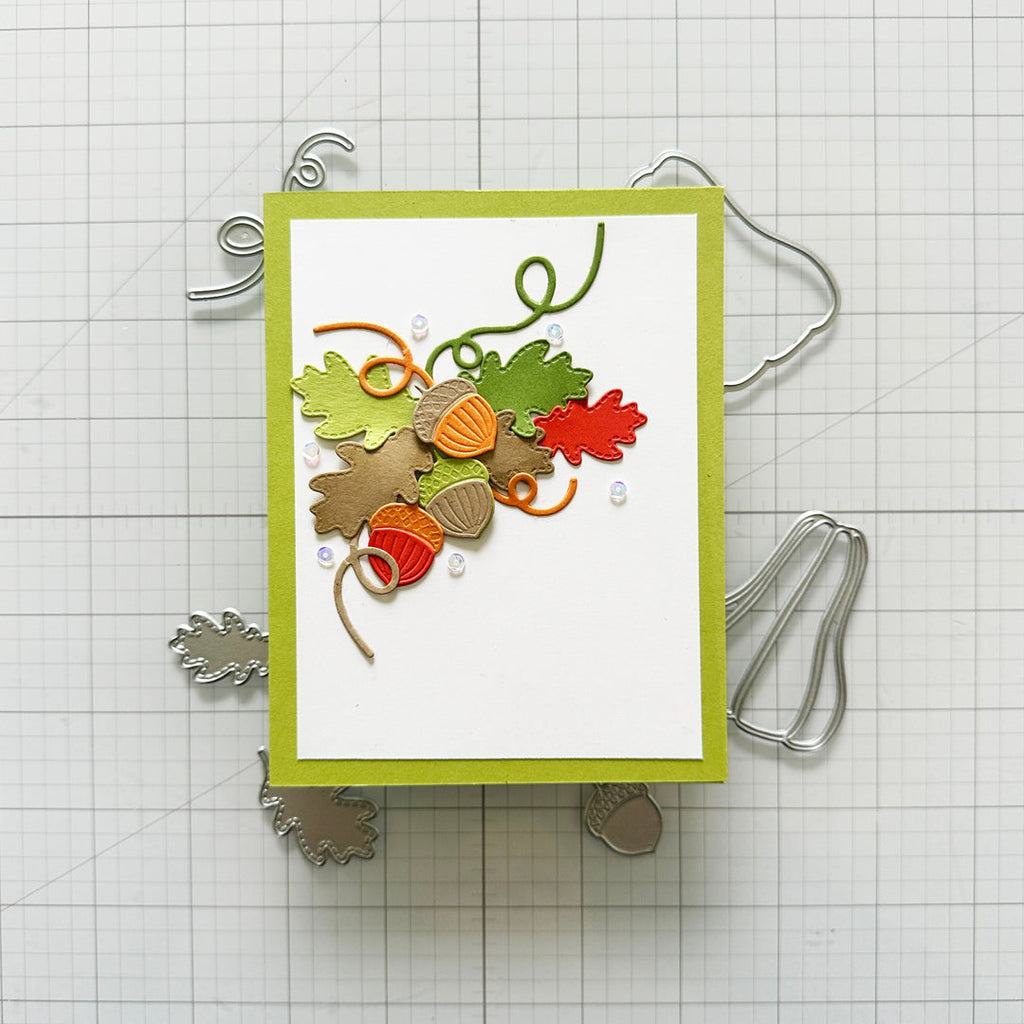 A handmade layered card featuring autumn-themed embellishments including green and orange leaves, acorns, and curled ribbon on a white background with a lime green border, displayed on a gridded craft mat with metal dies.