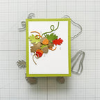 A handmade layered card featuring autumn-themed embellishments including green and orange leaves, acorns, and curled ribbon on a white background with a lime green border, displayed on a gridded craft mat with metal dies.