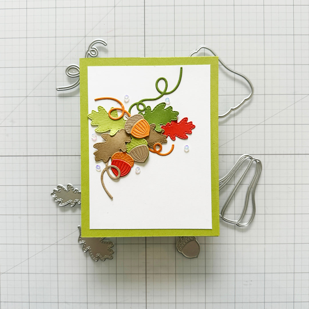 A handmade layered card featuring autumn-themed embellishments including green and orange leaves, acorns, and curled ribbon on a white background with a lime green border, displayed on a gridded craft mat with metal dies.