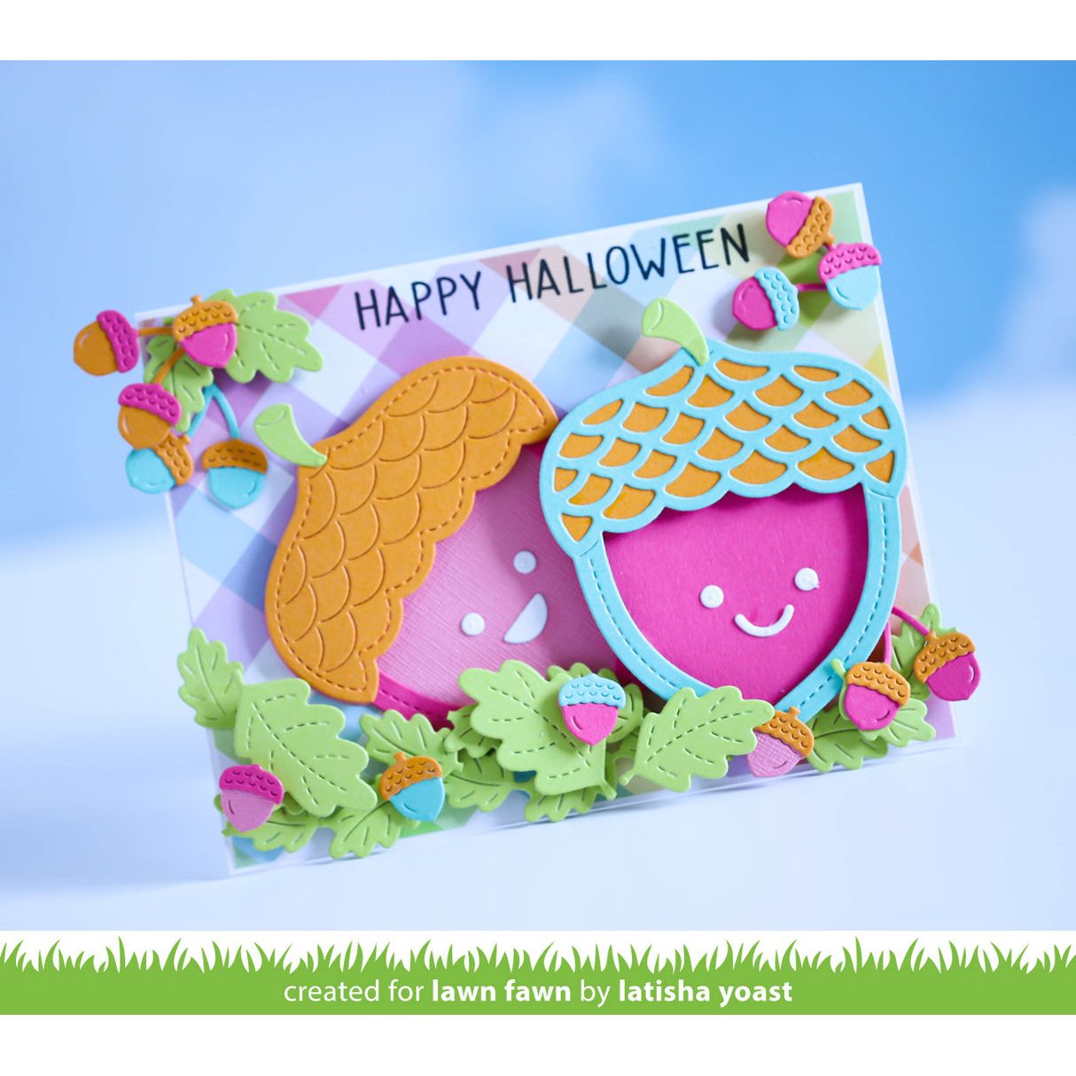 A handmade layered card featuring cute acorns and leaves with pink faces and colorful acorn details, with the text 'HAPPY HALLOWEEN' on a pastel plaid background. A green banner at the bottom reads 'created for lawn fawn by latisha yoast'.
