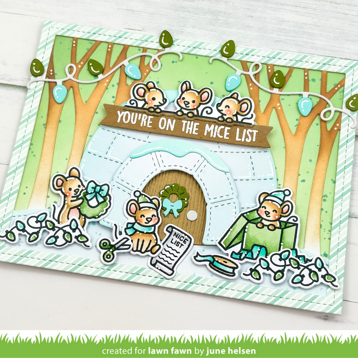 A handmade layered card featuring cute cartoon mice around a small white igloo with a wooden door and a brown banner reading 'YOU'RE ON THE MICE LIST'. The card includes green striped borders, string lights, trees, and small die-cut mouse decorations, photographed on a white wood surface.