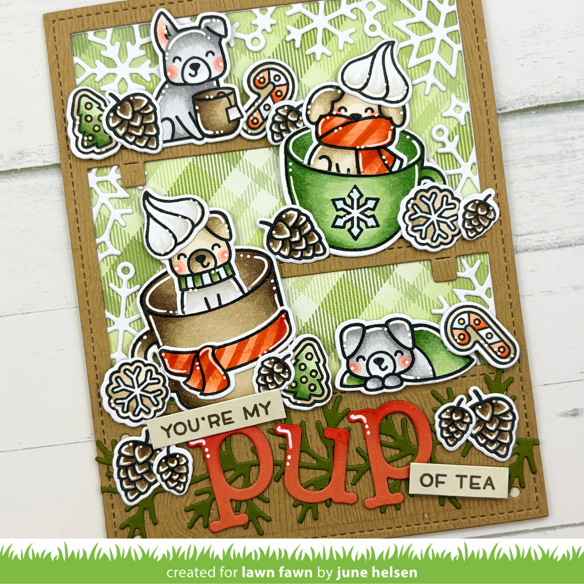 A handmade layered card featuring four illustrated puppy tea-time scenes with pinecones, snowflakes, candy canes, and the phrase 'YOU'RE MY pup OF TEA'. The card is photographed on a white wood background with a green grass-like strip at the bottom reading 'created for lawn fawn by june helsen'.