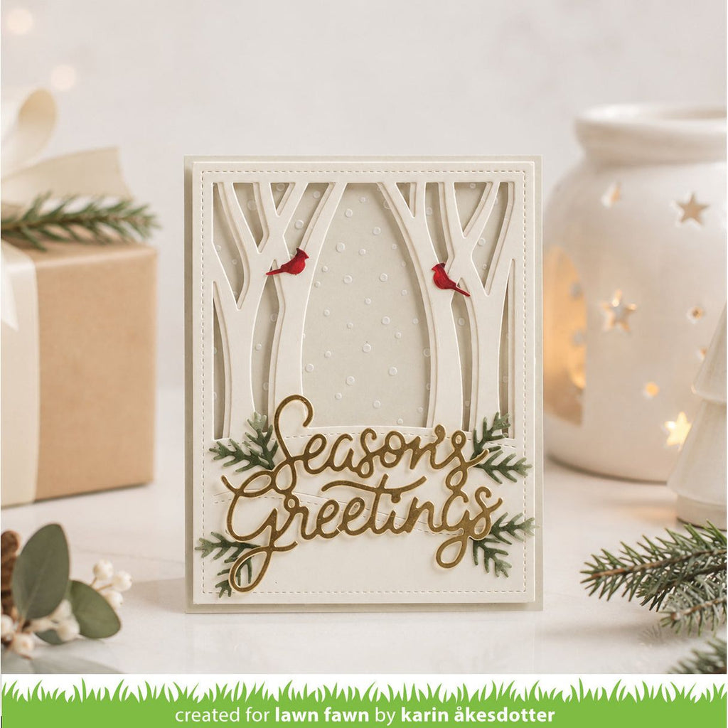 A handmade layered card with a forest theme, featuring laser-cut tree branches, two small red birds, green pine branches, and gold foil script reading 'Seasons Greetings'. The card is displayed on a white surface with a wrapped gift, eucalyptus sprigs, and a glowing white starry candle holder in the background.