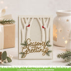 A handmade layered card with a forest theme, featuring laser-cut tree branches, two small red birds, green pine branches, and gold foil script reading 'Seasons Greetings'. The card is displayed on a white surface with a wrapped gift, eucalyptus sprigs, and a glowing white starry candle holder in the background.