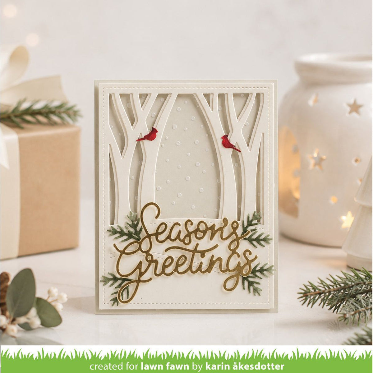 A handmade layered card with a forest theme, featuring laser-cut tree branches, two small red birds, green pine branches, and gold foil script reading 'Seasons Greetings'. The card is displayed on a white surface with a wrapped gift, eucalyptus sprigs, and a glowing white starry candle holder in the background.