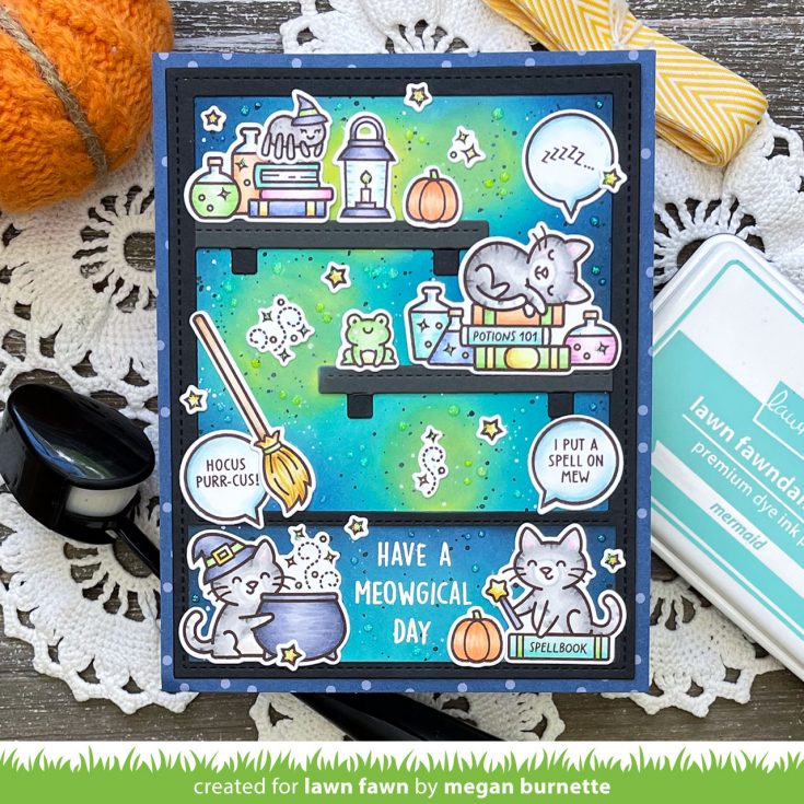 A handmade layered card with a spooky cat-themed design, displayed on a rustic wooden surface with knitting needles, yarn, and a stamp ink pad. The card features a blue and green gradient background with cartoon cats, potion bottles, a broom, and speech bubbles reading 'HAVE A MEOWGICAL DAY'. | color-code:alt2