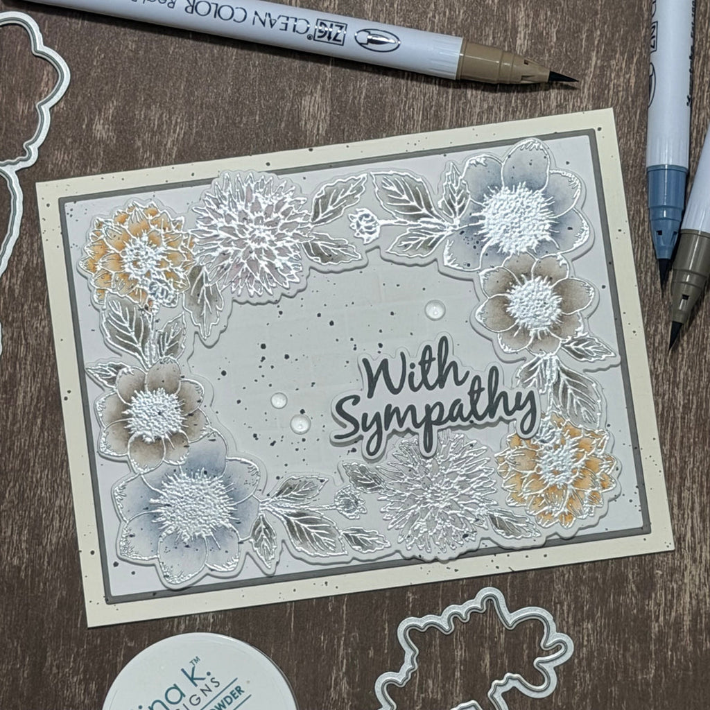 A handmade layered card with floral die-cut designs and the words 'With Sympathy' in black script, displayed on a wooden surface with crafting tools around it.