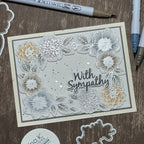 A handmade layered card with floral die-cut designs and the words 'With Sympathy' in black script, displayed on a wooden surface with crafting tools around it.