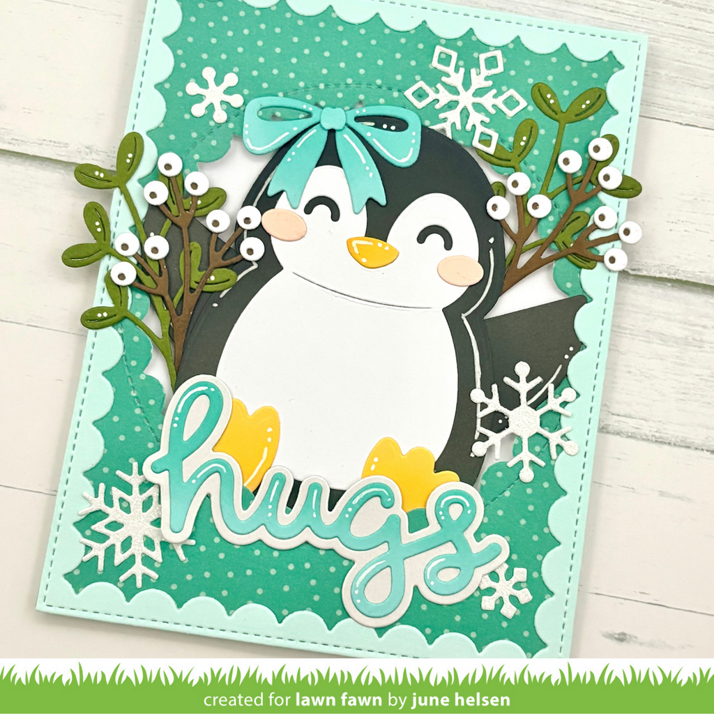 A handmade layered greeting card featuring a penguin with a teal bow, surrounded by greenery, white snowflakes, and the word 'hugs' in teal script. The card is photographed on a white wood background with a green grass banner at the bottom reading 'created for lawn fawn by june helsen'.