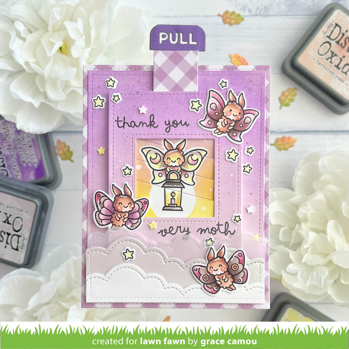 A handmade layered greeting card with butterfly and angel illustrations, shown on a styled flat-lay background with white flowers and ink pads. The card has a purple pull tab and reads 'thank you very moth'.