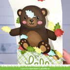A handmade layered paper card featuring a cute brown bear with colorful autumn leaves and acorns, held open by a hand. The card has a green and white plaid base with the text 'it's lawn fawn' at the bottom.