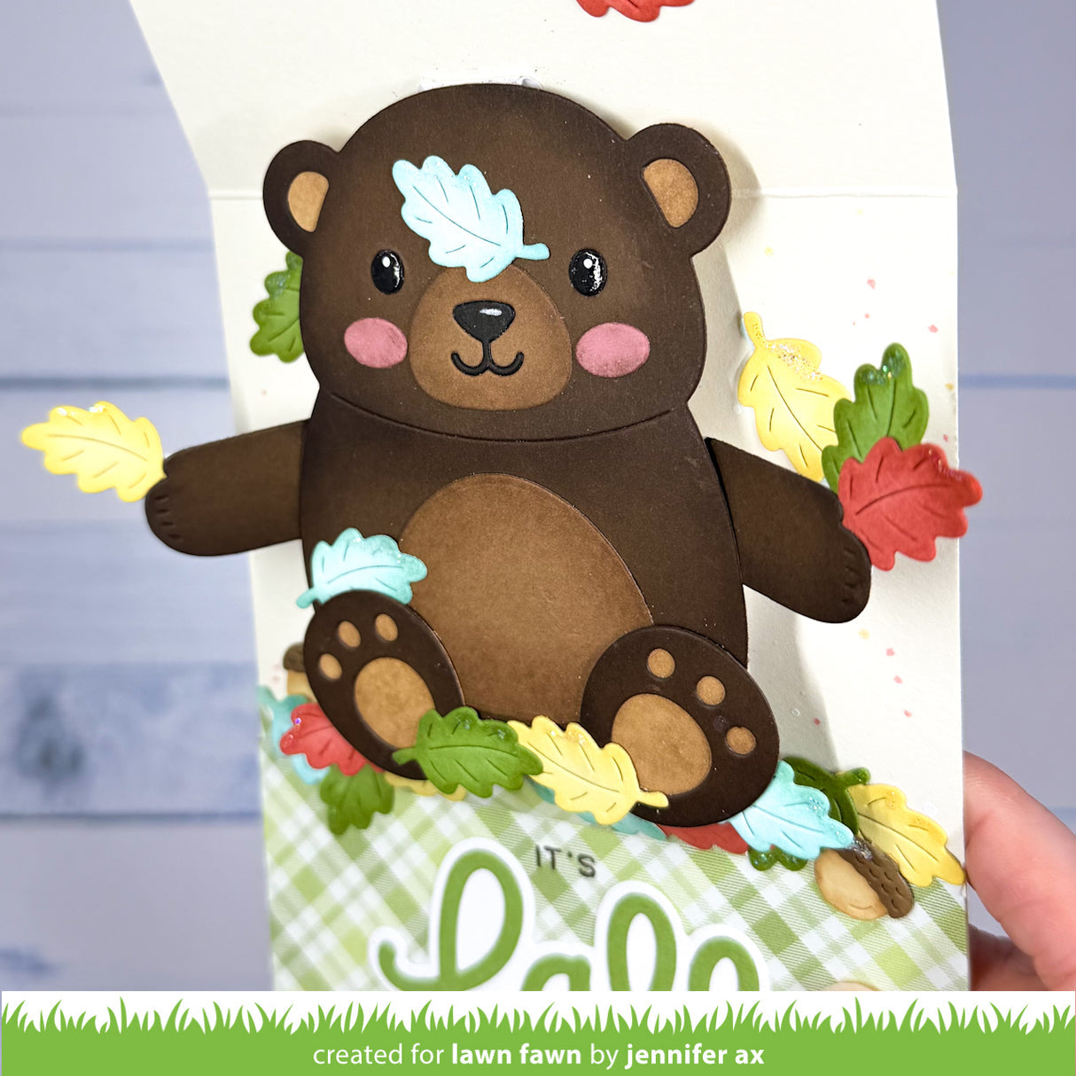 A handmade layered paper card featuring a cute brown bear with colorful autumn leaves and acorns, held open by a hand. The card has a green and white plaid base with the text 'it's lawn fawn' at the bottom.