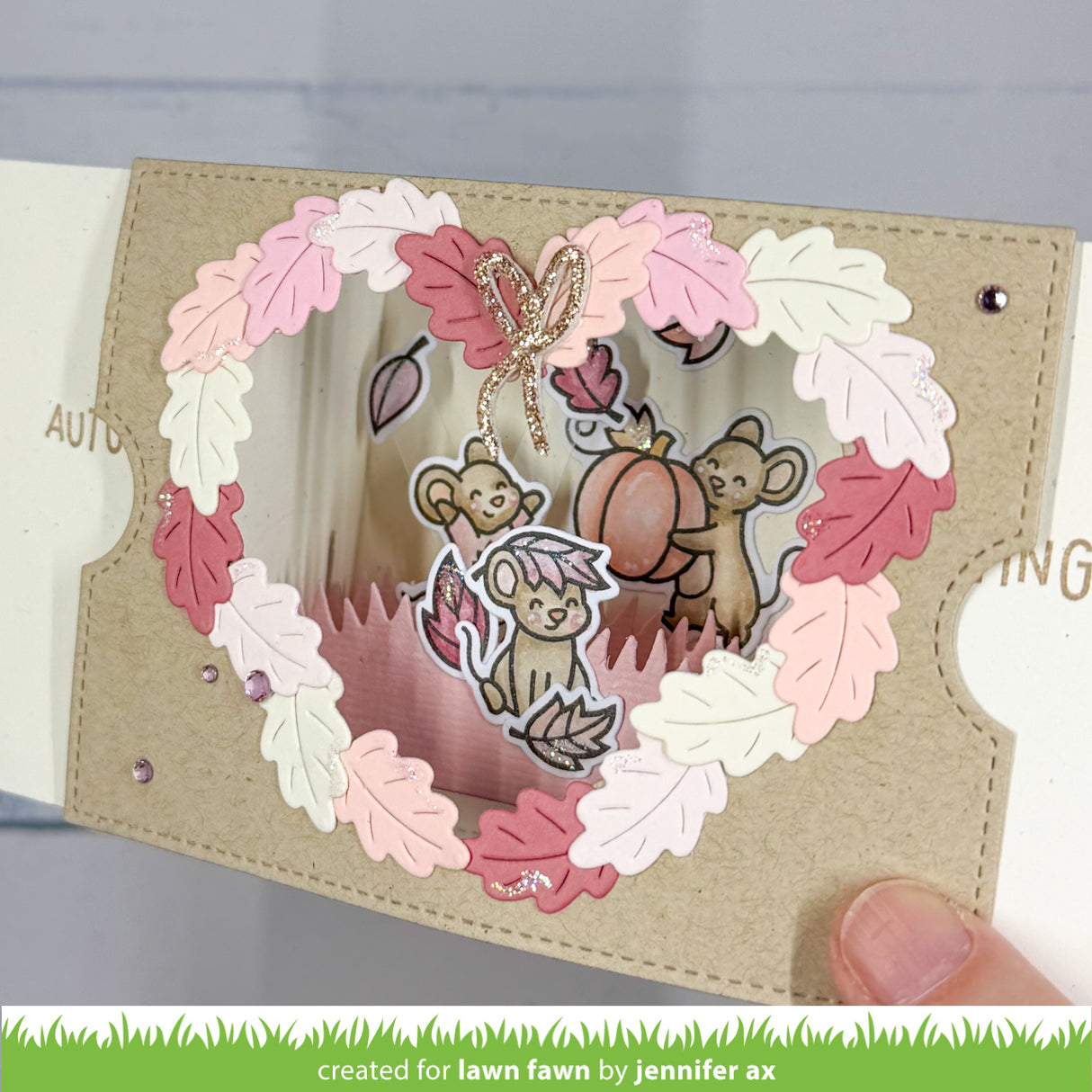 A handmade layered paper card with a circular autumn-themed design. The card features a beige textured paper border, a wreath of pink, white, and brown paper leaves, and a central pop-up window showing  a mouse, a squirrel, and a pumpkin.