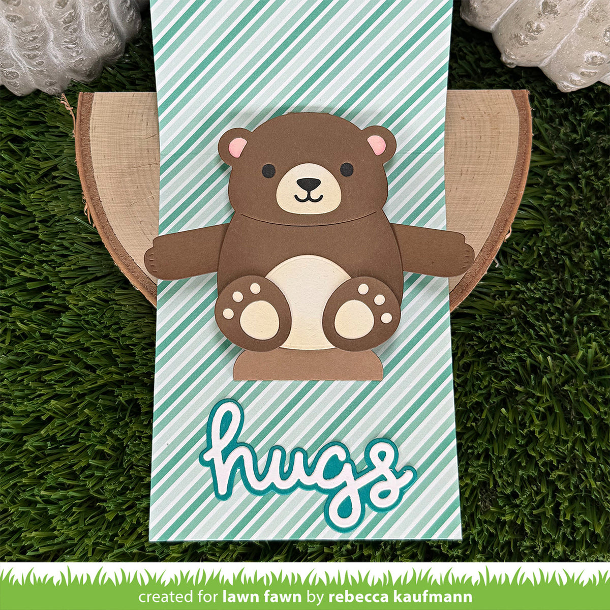 A handmade paper craft card featuring a cute brown bear with a cream snout and paws, set against a mint green and white striped background with the word 'hugs' in white script. 