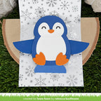 A handmade penguin card or decoration made from layered blue, white, and orange paper, displayed on a patterned white sheet over a wood slice and artificial green grass. A green banner at the bottom reads 'created for lawn fawn by rebecca kaufmann'.