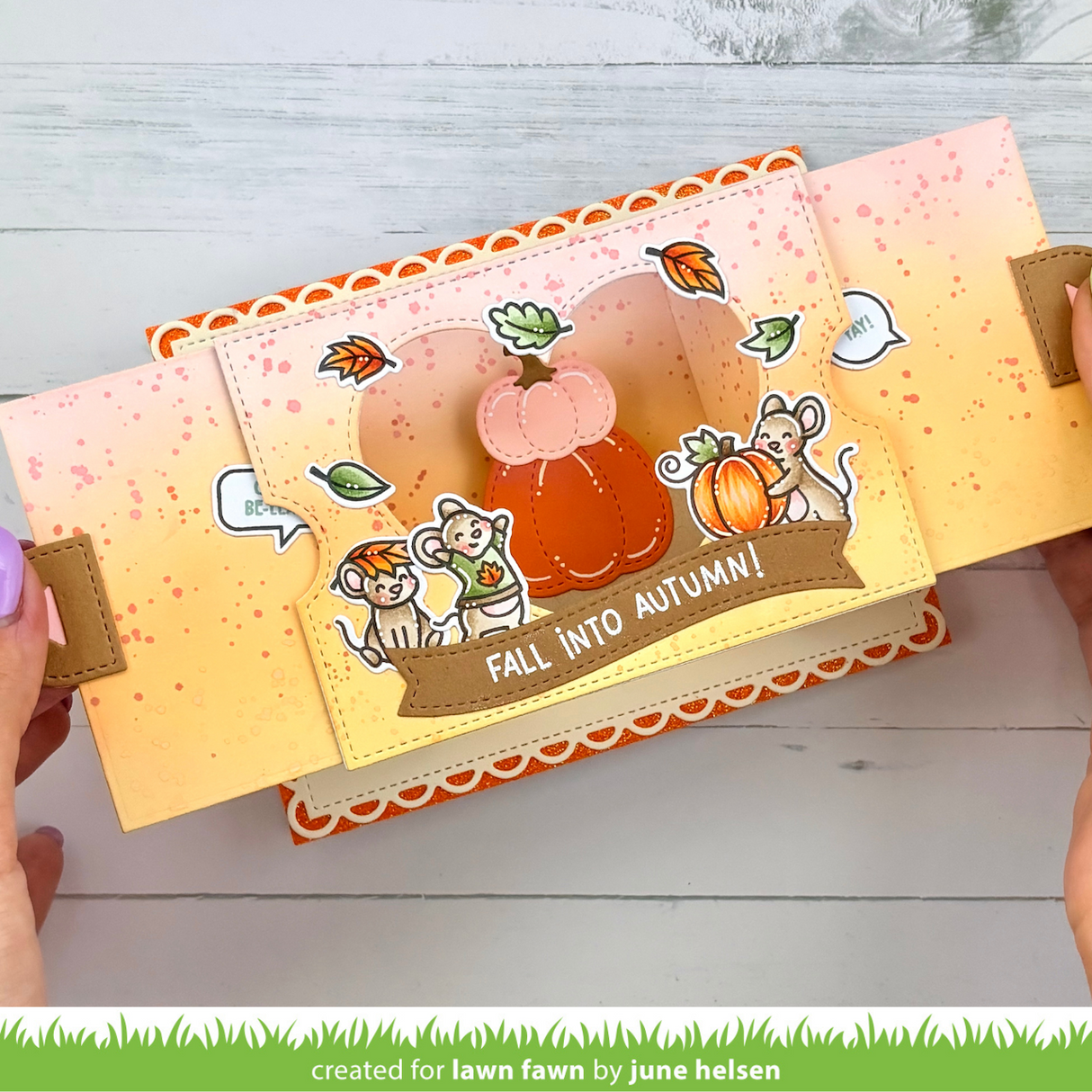 A handmade pop-up card art piece with autumn themes, held open by two hands. The design features a 3D pumpkin, cartoon mice, fall leaves, and the text 'FALL INTO AUTUMN!' on a warm orange and yellow gradient background with speckles and scalloped trim.