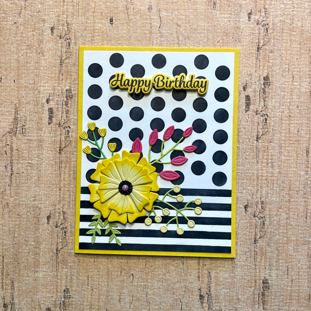 A handmade square birthday card with a yellow border, featuring a black and white polka dot pattern, black and white horizontal stripes, a large yellow flower, pink flower buds, and 'Happy Birthday' in yellow script. The card is displayed on a textured light brown wood background.
