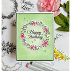 A handmade square greeting card with a light green background, featuring a stamped floral wreath and the words 'Happy Birthday' in black script. The card is styled in a flat-lay arrangement with pink and yellow flowers, green leaves, and crafting tools on a white surface.