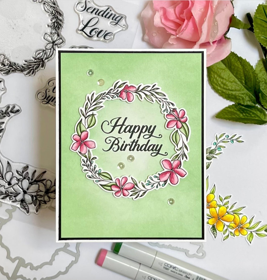 A handmade square greeting card with a light green background, featuring a stamped floral wreath and the words 'Happy Birthday' in black script. The card is styled in a flat-lay arrangement with pink and yellow flowers, green leaves, and crafting tools on a white surface.