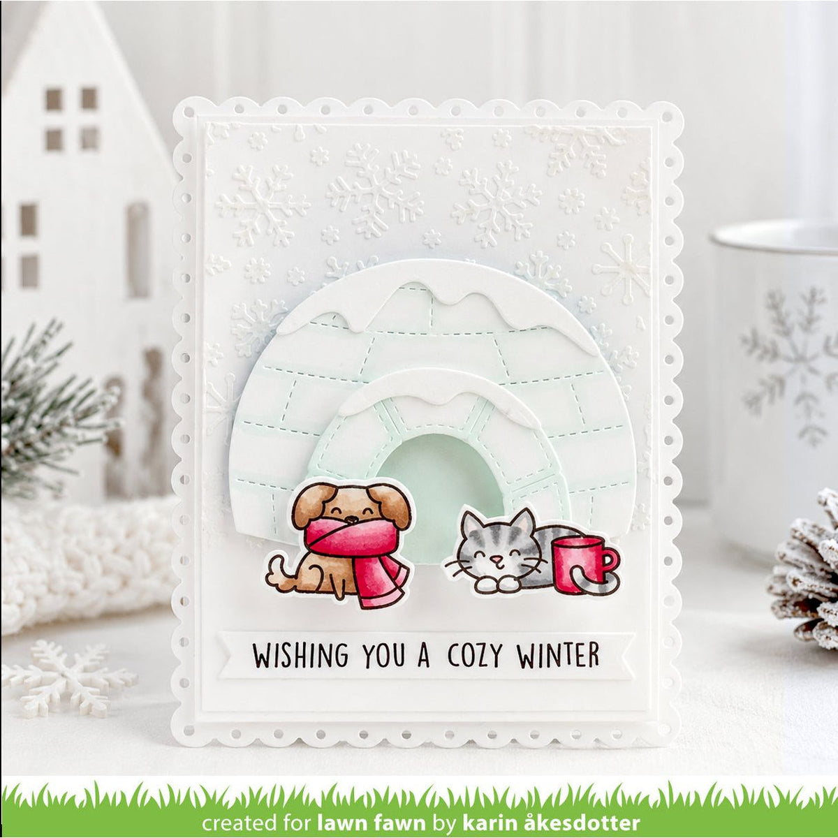 A handmade square greeting card with a scalloped white border, featuring a winter scene with a dog in a pink scarf, a gray cat, and a red mug. The card reads 'WISHING YOU A COZY WINTER' and is displayed in a styled holiday setting.