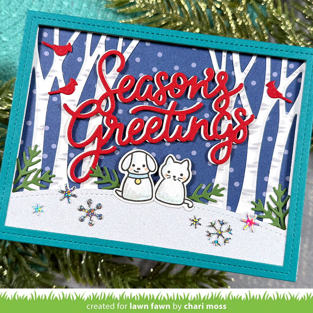 A handmade square greeting card with a winter forest scene, featuring the words 'Season's Greetings' in red script, two small white dog and cat figures, red birds, and snowflake embellishments. The card is styled with green pine branches in the background.
