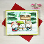 A handmade winter greeting card featuring three cute snowman characters in colorful hats and scarves holding mugs, with a sign that reads 'STAY COZY'. The card is styled on top of red envelopes on a light wood background, with 'the Greeting Farm' branding visible.