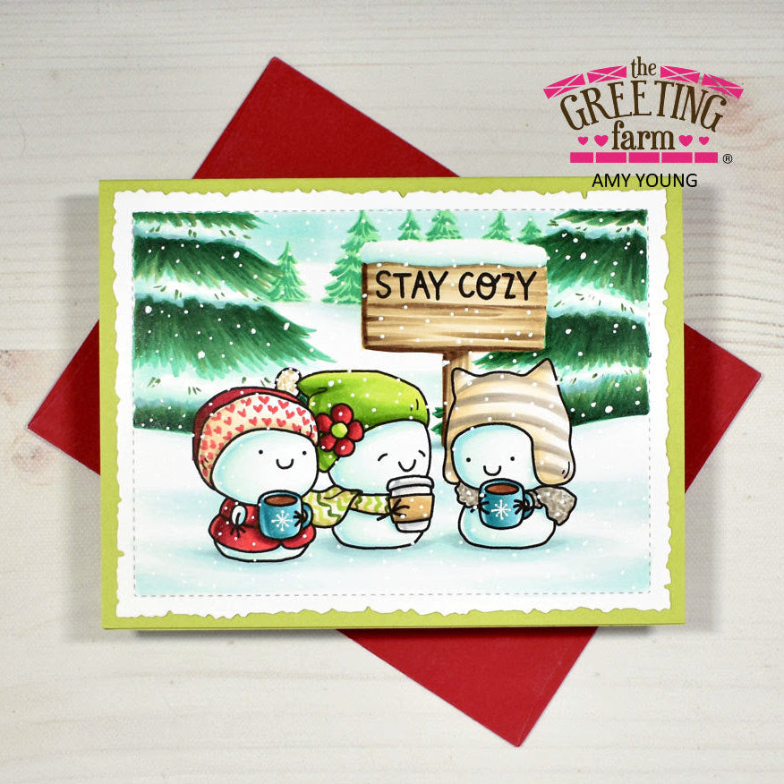 A handmade winter greeting card featuring three cute snowman characters in colorful hats and scarves holding mugs, with a sign that reads 'STAY COZY'. The card is styled on top of red envelopes on a light wood background, with 'the Greeting Farm' branding visible.