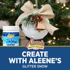 A jar of Aleene's Glitter Snow is shown next to a decorated white Christmas ornament with a burlap bow, pinecone, and gold bell, set against a blurred Christmas tree background. A blue banner at the bottom reads 'CREATE WITH ALEENE'S GLITTER SNOW' with the Aleene's logo.