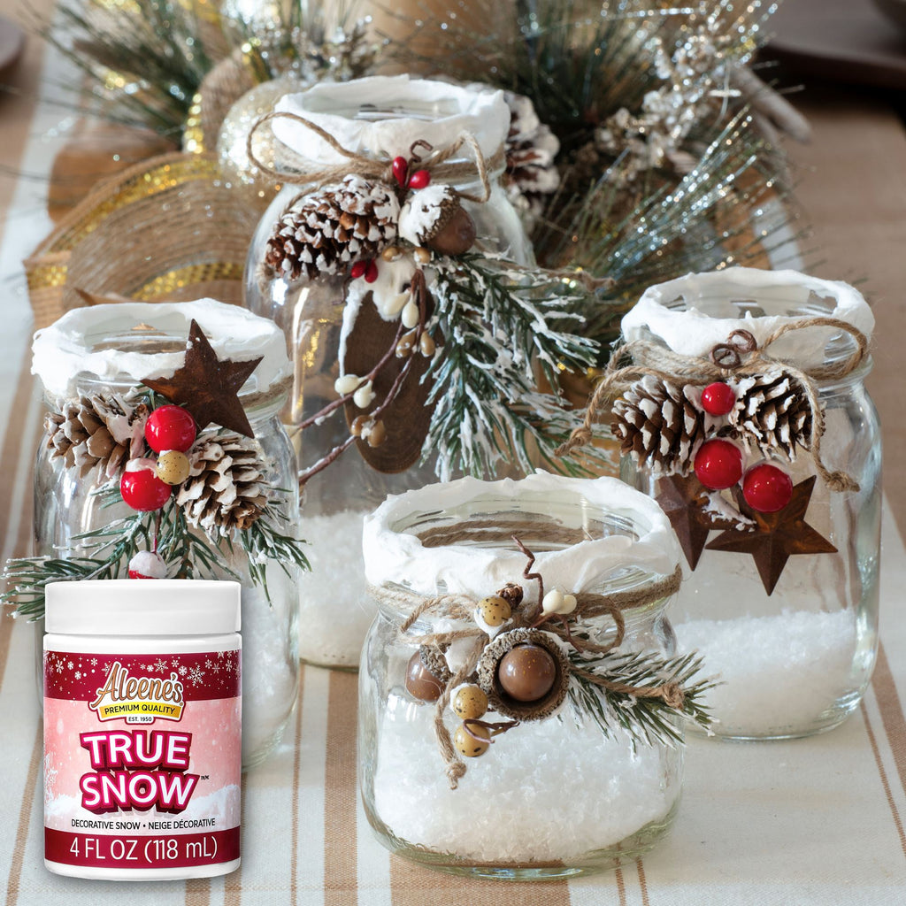 A jar of Aleene's True Snow Non-Toxic Glue shown next to several decorated glass jars filled with white faux snow, adorned with pinecones, evergreen branches, burlap ribbon, and red berries.