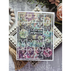 A rectangular jigsaw puzzle card with a floral and abstract multicolor pattern, featuring a white border and the words 'Love you to pieces' in the center. The card is styled on a gray textured surface with decorative props including a tassel, wooden beads, a pink rose, and a small tray.