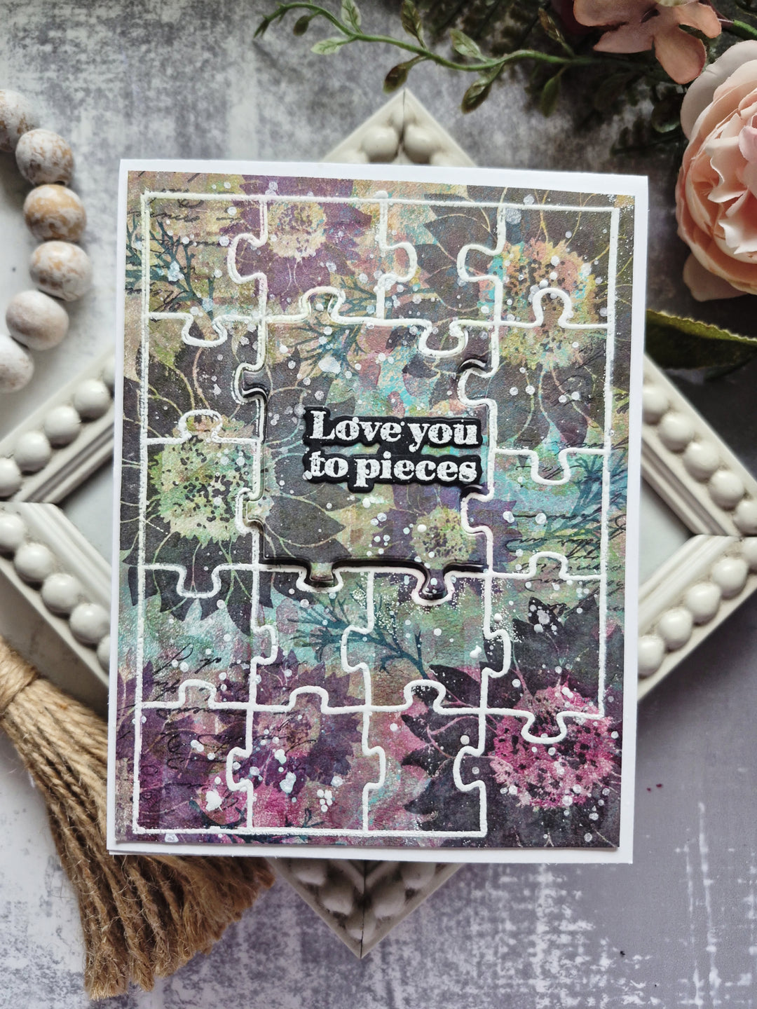A rectangular jigsaw puzzle card with a floral and abstract multicolor pattern, featuring a white border and the words 'Love you to pieces' in the center. The card is styled on a gray textured surface with decorative props including a tassel, wooden beads, a pink rose, and a small tray.