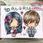 A square printed card or stamp design featuring chibi-style cartoon couple characters with the text 'Mr & Mrs' and 'I DO', plus hearts and a ring. The card is photographed on a light wood surface with decorative elements around it.