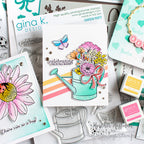 A styled flat-lay of Gina K. Design stamps and cards, featuring a central white card with a colorful watering can and flower bouquet illustration, surrounded by other stamped and printed craft pieces.