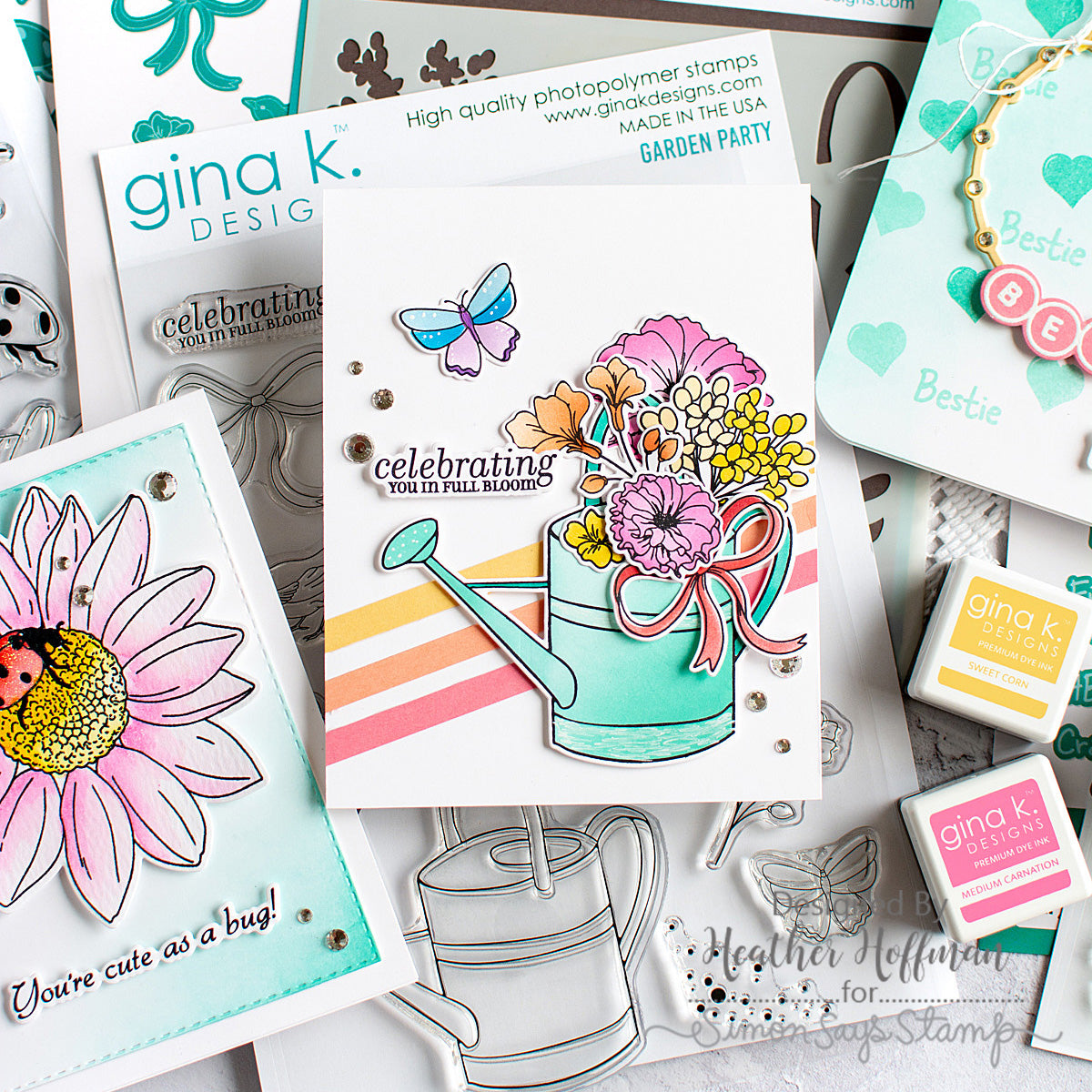A styled flat-lay of Gina K. Design stamps and cards, featuring a central white card with a colorful watering can and flower bouquet illustration, surrounded by other stamped and printed craft pieces.