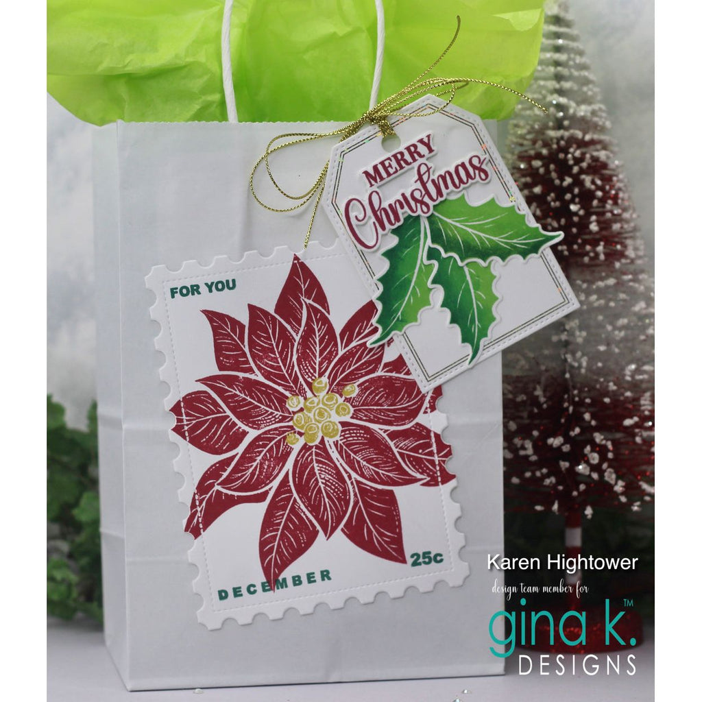 A white gift bag with a green tissue top and a gold ribbon, featuring a die-cut postage stamp design of a red poinsettia with a hanging 'Merry Christmas' tag. The photo is branded with 'gina k.