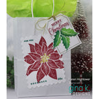 A white gift bag with a green tissue top and a gold ribbon, featuring a die-cut postage stamp design of a red poinsettia with a hanging 'Merry Christmas' tag. The photo is branded with 'gina k.